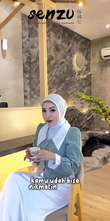 evimonikaa duduk memegang cangkir di depan dinding batu dan bonsai di Senzu Reflexology